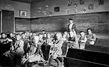 Primary class in the Highlandlake school about 1880. Their teacher is standing at the back of the room while the children are seated at t heir school desks. Pictures from the era or on the wall above the painted, wood blackboards.