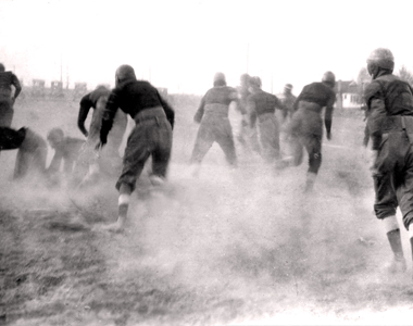 Football 1921 style. The boys are scrimaging in a field next to the school. dust if flying, and the football is in play.