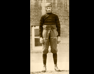 1921, boy in football uniform. Leather hat, jersy shirt and a highwaisted pants that go to the knee. There is padding on the legs and a wide belt around the waist.