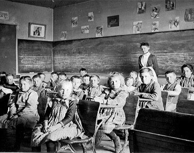 Highlandlake schoolroom circa 1883. The chuildren are sitting in their seats while their teacher is standing at the back of the room. All ages are represented.